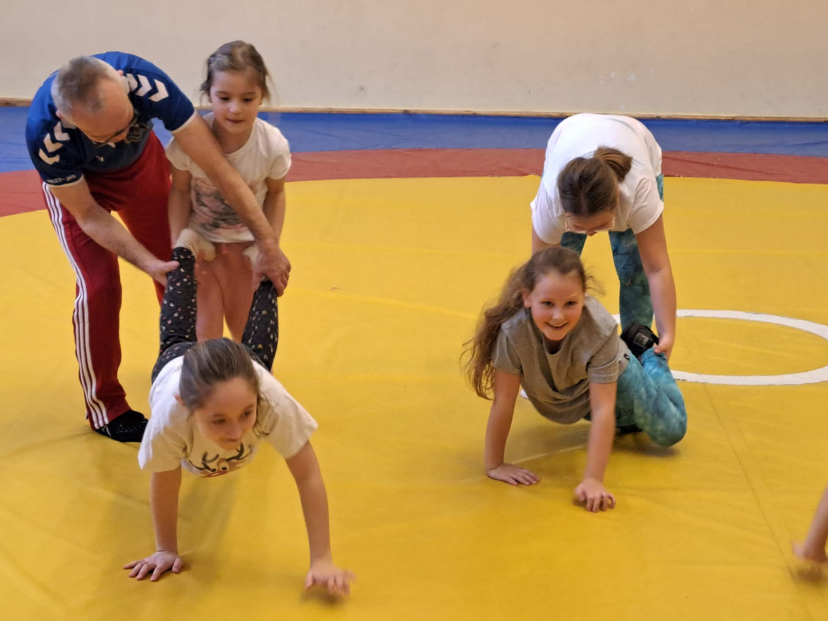Dzieci na sali gimnastycznej opierają się o podłogę na rękach a ludzie trzymają je za nogi