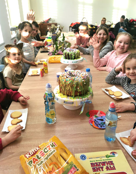 Przy stole zastawionym ozdobami wielkanocnymi oraz ciastami siedzą dzieci, po obu stronach