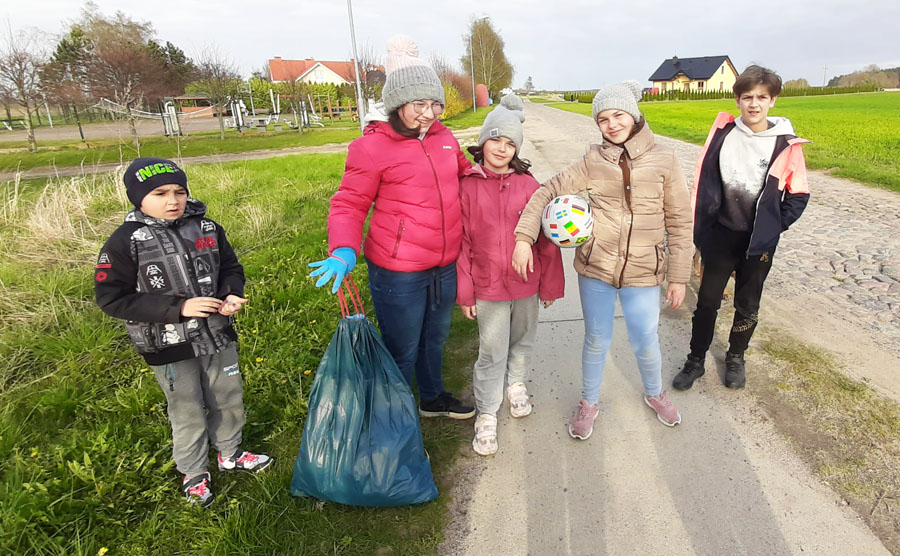 Pięcioro dzieci, 3 dziewczynki i dwóch chłopców, stoją obok siebie na chodniku przy wiejskiej drodze, z jednym workiem na śmieci, w tle pole i dwa domki jednorodzinne