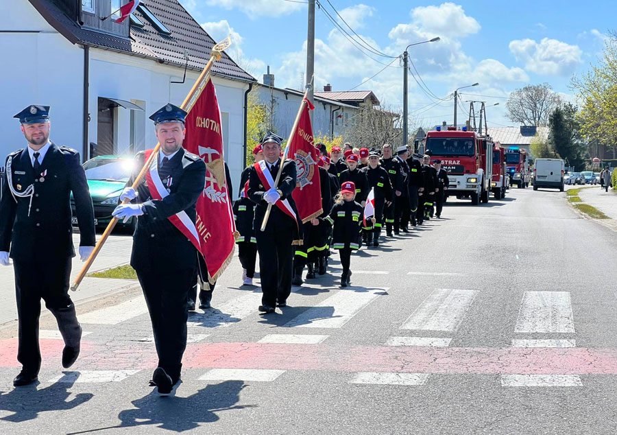 Na ulicy idzie pochód, na przedzie dwóch strażaków w mundurach galowych, jeden z flagą, za nimi podobna para, za nimi strażacy w różnych mudnurach, a za nimi jadą wozy strażackie