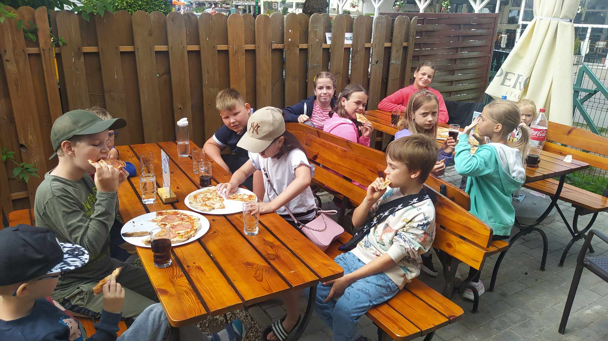 Na zewnątrz przy stoliku siedzą dzieci i jedzą pizzę.
