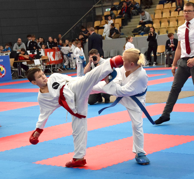 Dwóch zawodników karate walczy na macie, w tle ludzie