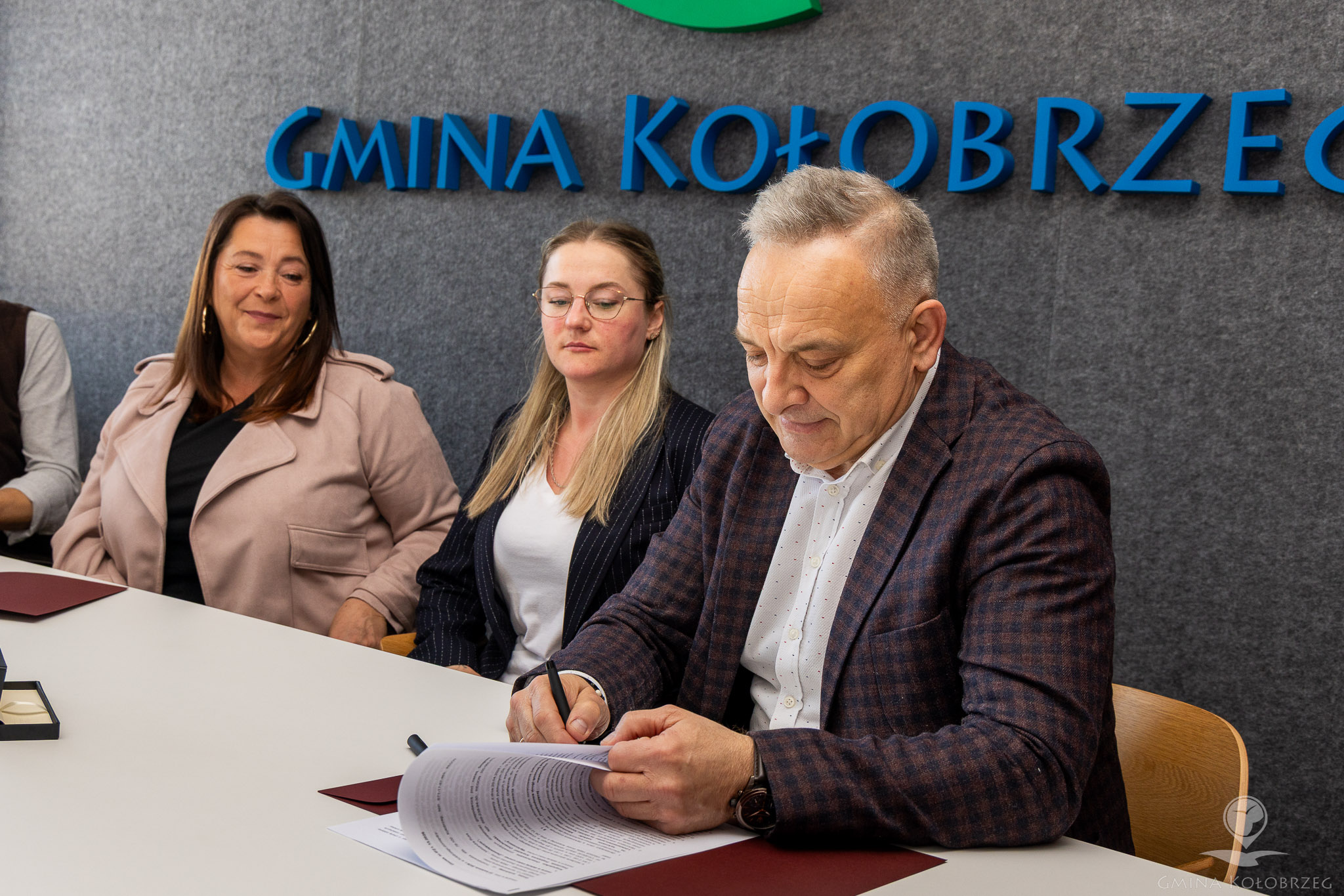 Na zdjęciu widać mężczyznę podpisującego dokument przy stole, na tle napisu &bdquo;Gmina Kołobrzeg&rdquo;. Obok siedzą dwie kobiety obserwujące sytuację. Ujęcie ma charakter oficjalny i przedstawia moment zawierania umowy.