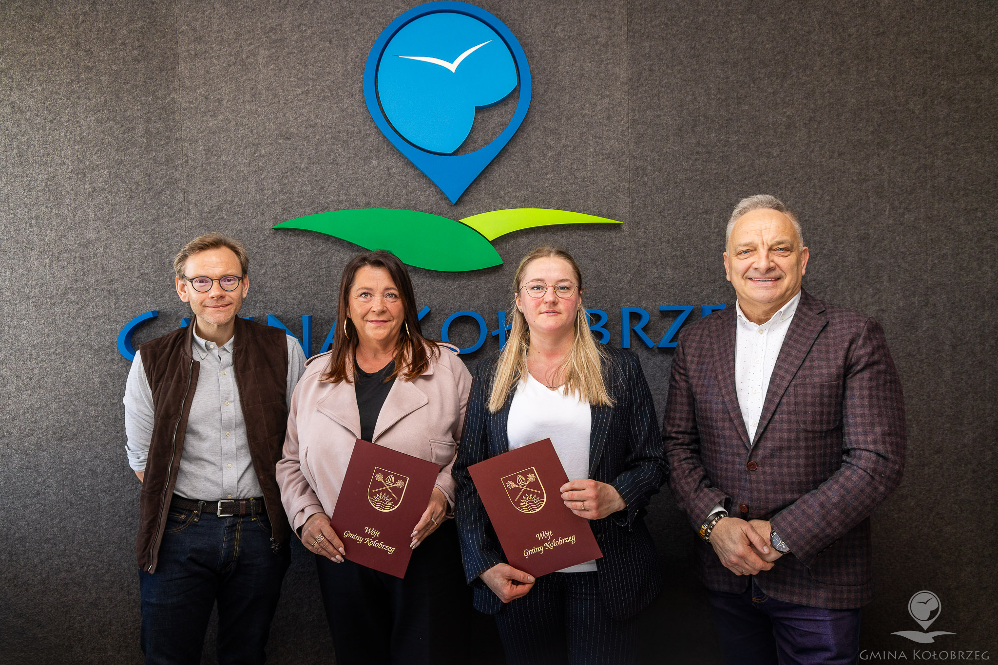 Na zdjęciu widać cztery osoby stojące na tle ściany z logo &bdquo;Gmina Kołobrzeg&rdquo;. Dwie kobiety w środku trzymają dokumenty, a obok stoją dwaj mężczyźni. Ujęcie ma charakter oficjalny i dokumentuje ważne wydarzenie, podpisanie umowy.
