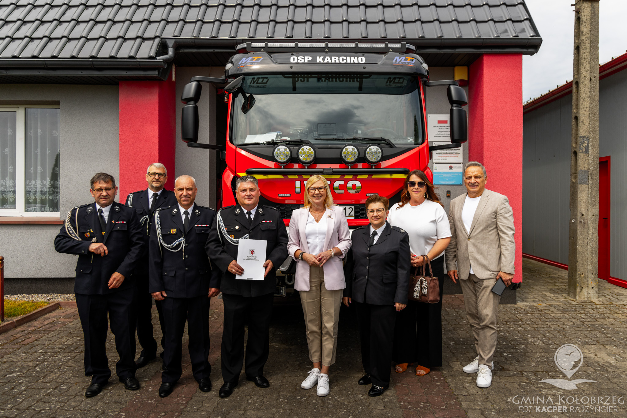 Zdjęcie przedstawiające strażak&oacute;w oraz przedstawicieli samorządu i lokalnej społeczności pozujących przed remizą Ochotniczej Straży Pożarnej w Karcinie. W tle widoczny jest nowoczesny w&oacute;z strażacki zaparkowany przed budynkiem jednostki.