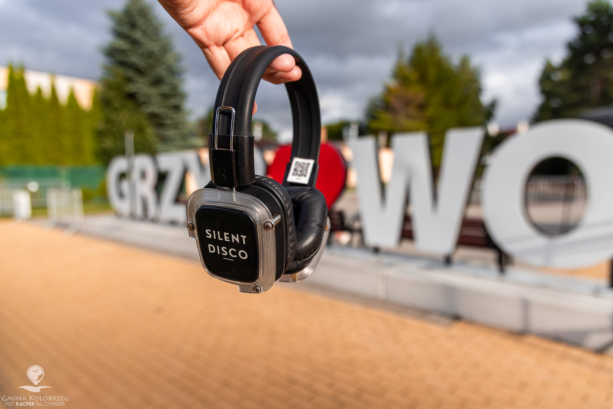 Zdjęcie słuchawek na tle napisu Zdjęcie przedstawia słuchawki z napisem „Silent Disco” trzymane na tle napisu „Grzybowo”. Ujęcie nawiązuje do plenerowego wydarzenia muzycznego, z rozmytym tłem i ciepłym światłem.